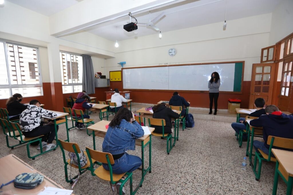 A classroom with students sitting at desks and a teacher standing in front of the chalkboard.