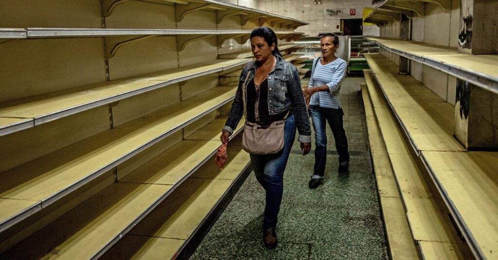 Two women are walking down a long aisle of empty shelves.