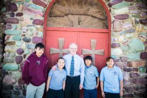 Four students and a teacher standing in front of a red double door with a wooden wheel design, set in a colorful stone wall.